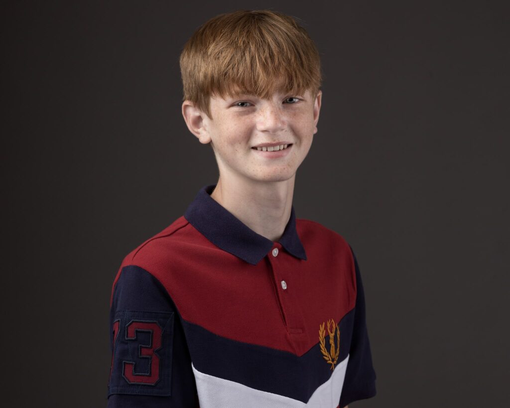 A young boy with reddish-brown hair smiling at the camera, wearing a red and navy polo shirt with a number 3 on the sleeve.