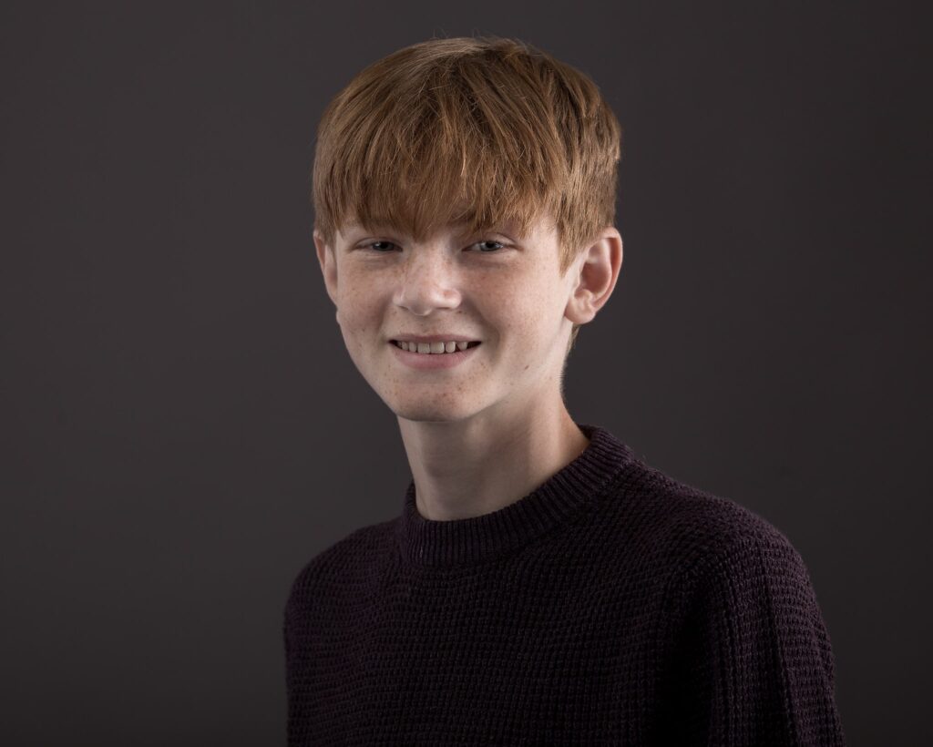 A young boy with reddish-brown hair and freckles smiles at the camera, wearing a dark knitted jumper against a grey background.