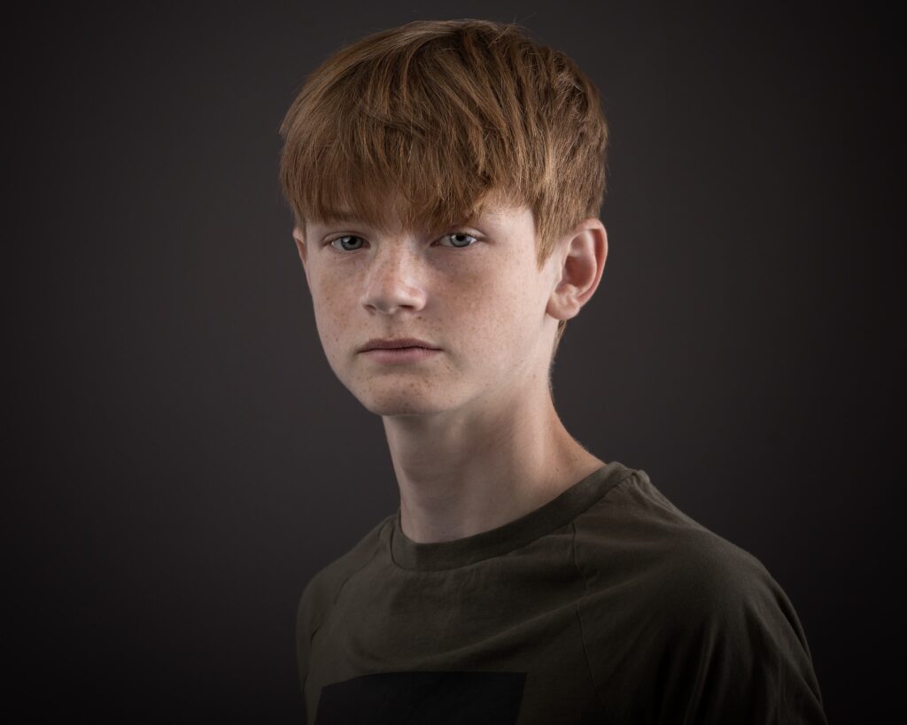 A young person with reddish-brown hair and freckles wearing a dark olive shirt against a dark grey background.