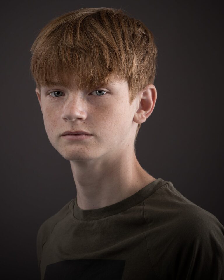 A young person with reddish-brown hair and freckles wearing a dark olive t-shirt, photographed against a grey background.