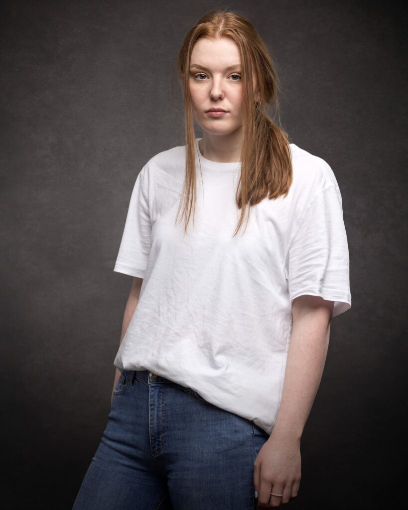 A young woman with long reddish-brown hair wearing a white t-shirt and blue jeans stands against a grey background.