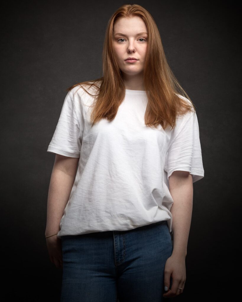 A young woman with long reddish-brown hair wearing a white t-shirt and blue jeans stands against a dark grey background.