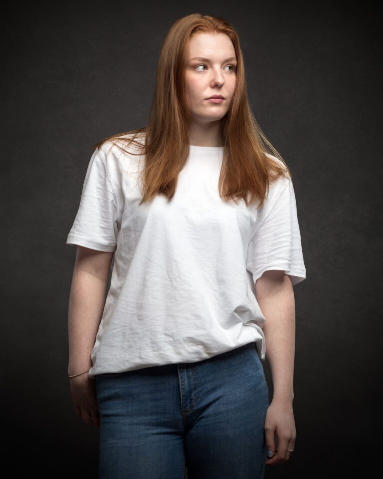 A young woman with long auburn hair wearing a white t-shirt and blue jeans stands against a dark grey background, looking off to the side.