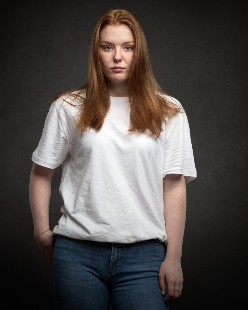 A person with long auburn hair wearing a white t-shirt and blue jeans stands against a dark grey background, looking toward the camera.