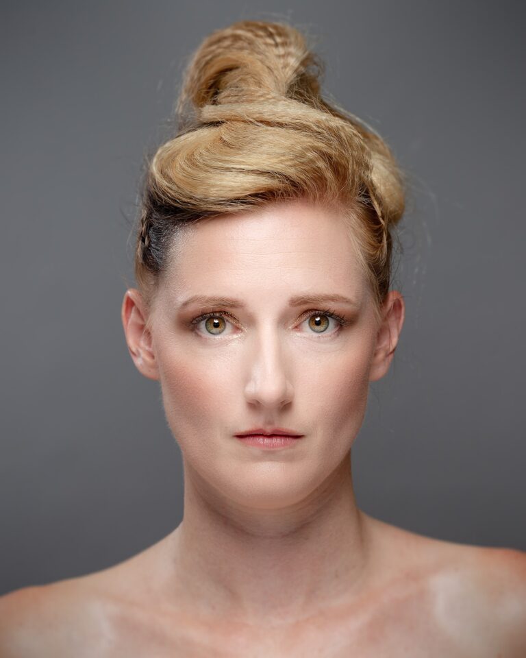 Woman with blonde hair styled in an updo, looking directly at camera against a grey background.