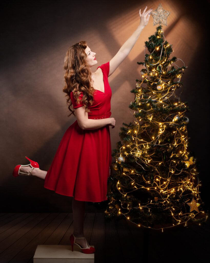 Woman in red dress placing a star on top of an illuminated Christmas tree against a brown backdrop.