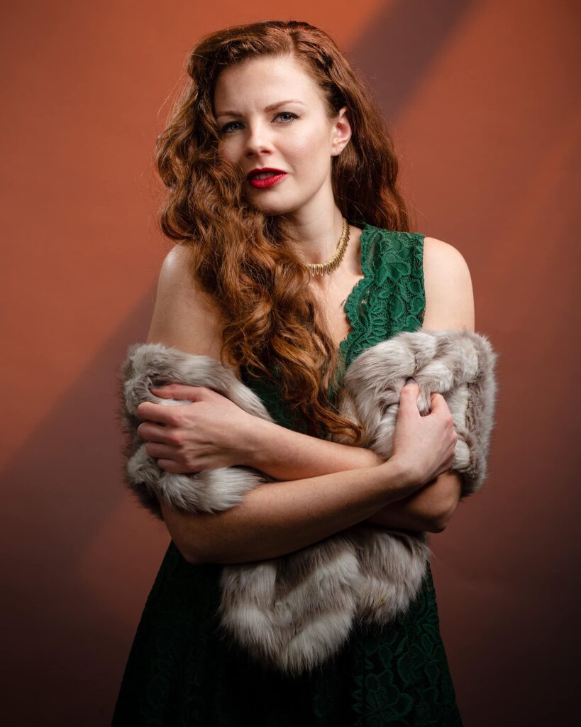 A woman with long wavy auburn hair and red lipstick wears a green lace top and grey fur stole against a warm orange background.
