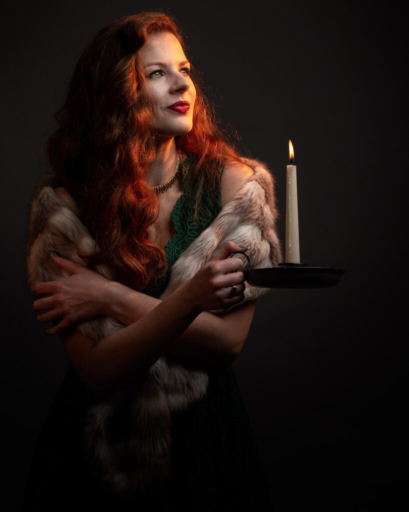 A woman with long reddish hair holds a candleholder with a lit candle, wearing fur and dark clothing against a black background.