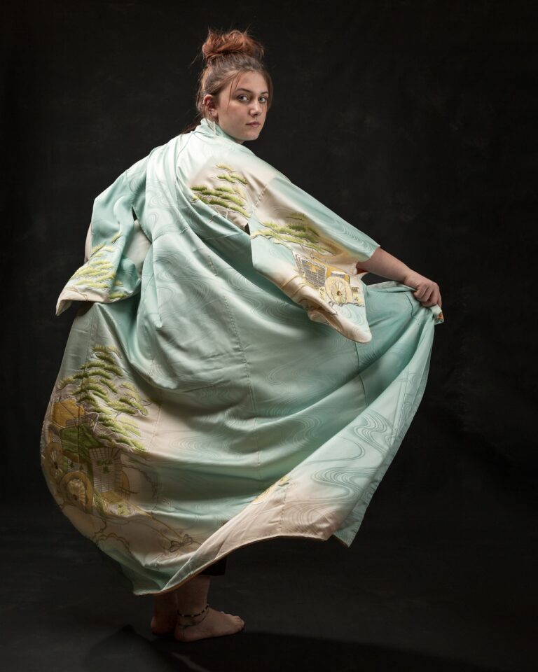 A woman wearing a flowing sage green silk robe with gold embroidered details, standing against a black background, looking over her shoulder.