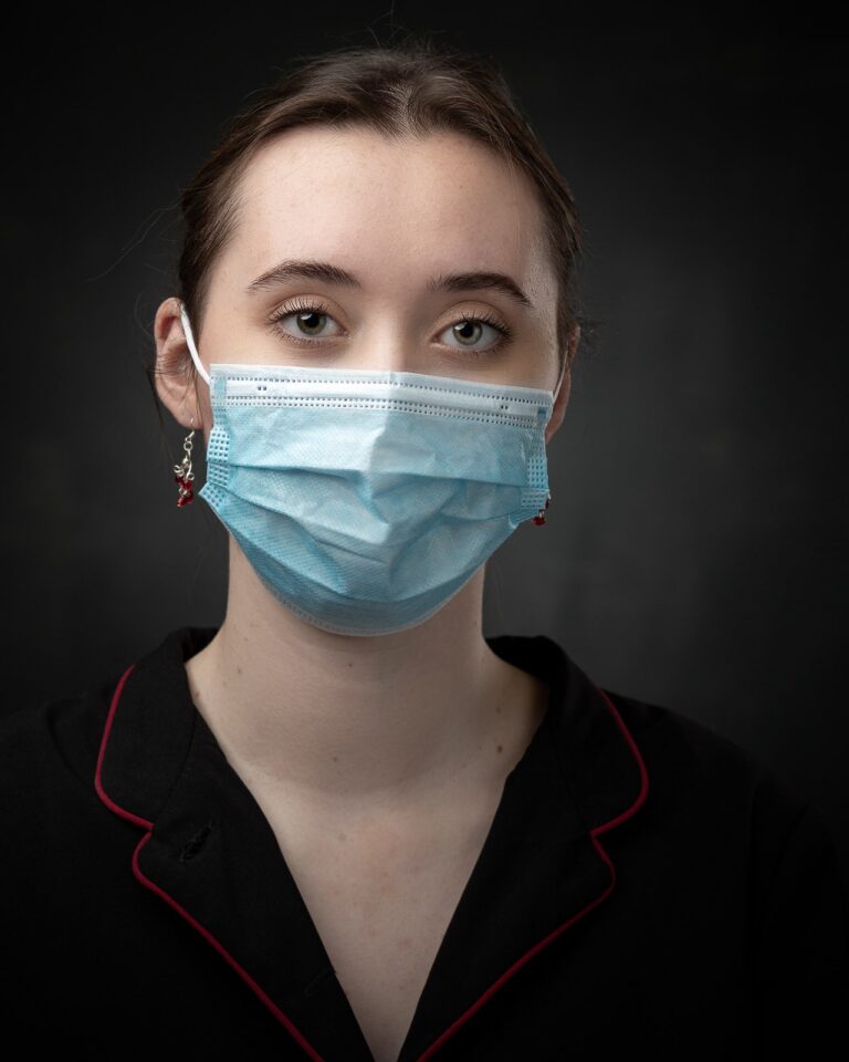 Woman wearing a blue surgical mask and dark jacket with red trim, against a dark background, looking directly at camera.