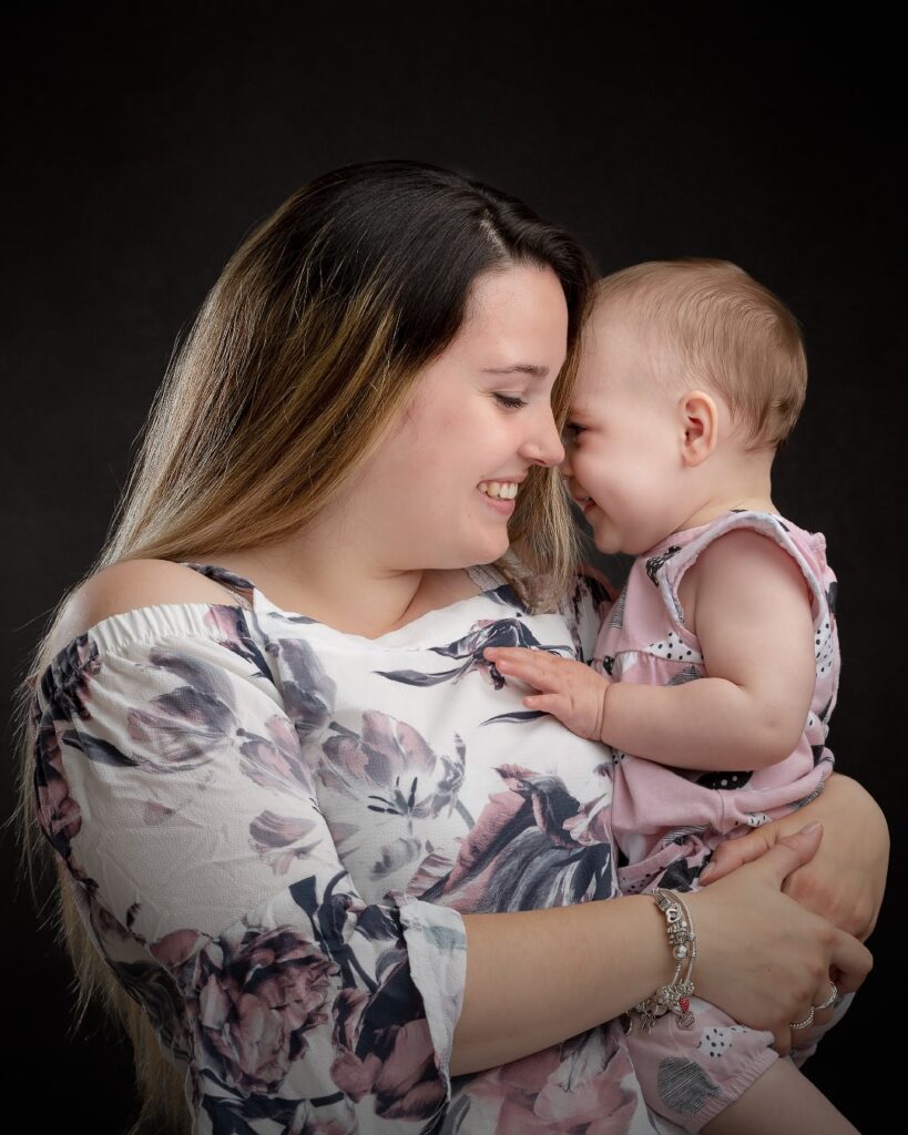 A woman holding a toddler, both smiling at each other against a black background. They wear floral patterned clothing.