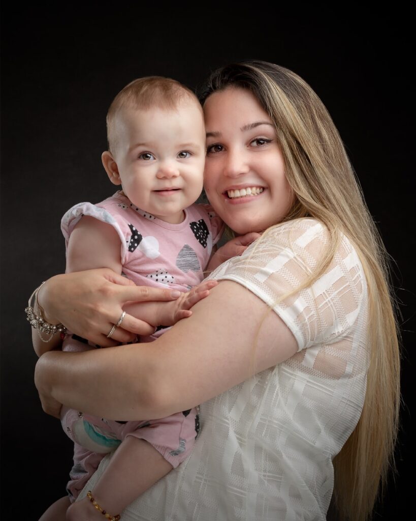 A woman with long blonde hair holds a toddler in a pink patterned top against a black background. Both are smiling at the camera.