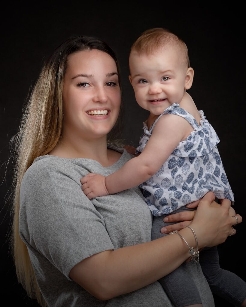 A woman holding a smiling toddler, both looking at the camera against a black background. Professional portrait.