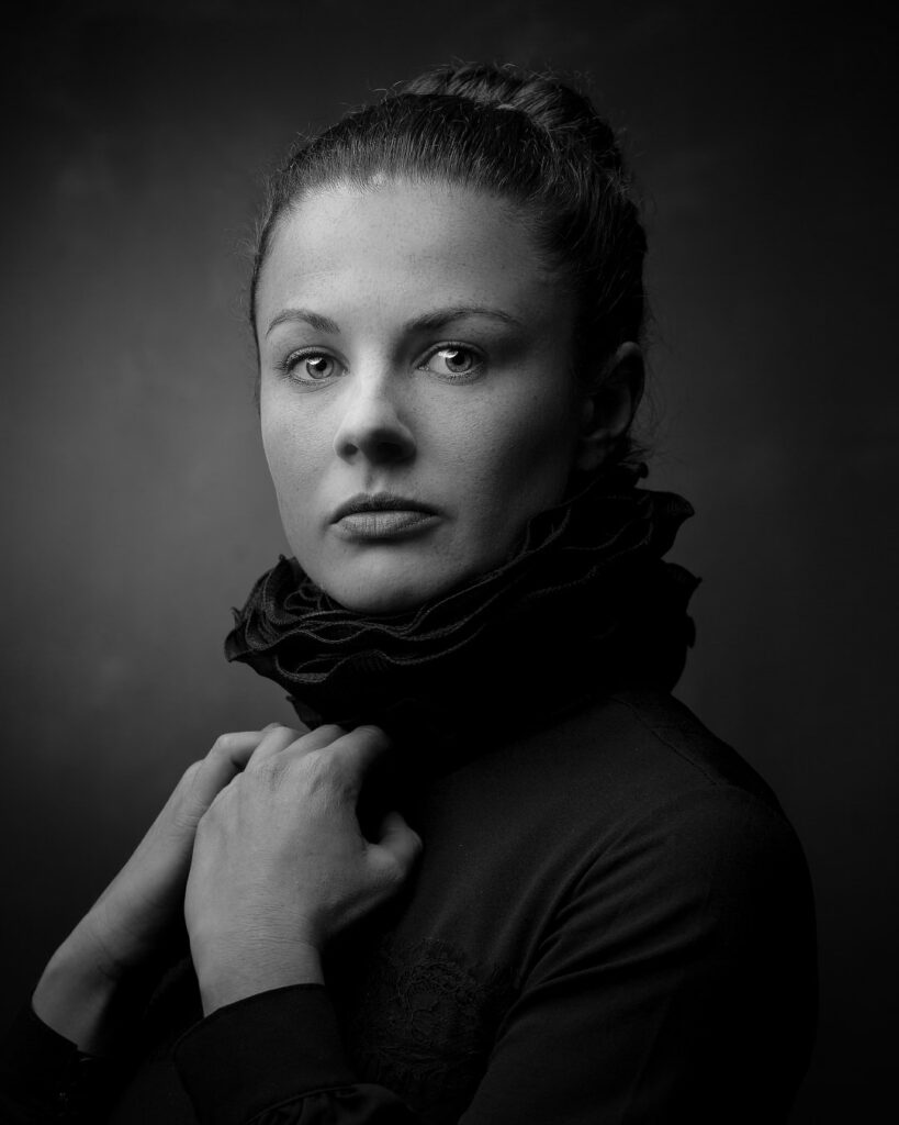 Black and white portrait of a woman with hair in a bun, wearing dark clothing with a scarf, against a grey background.