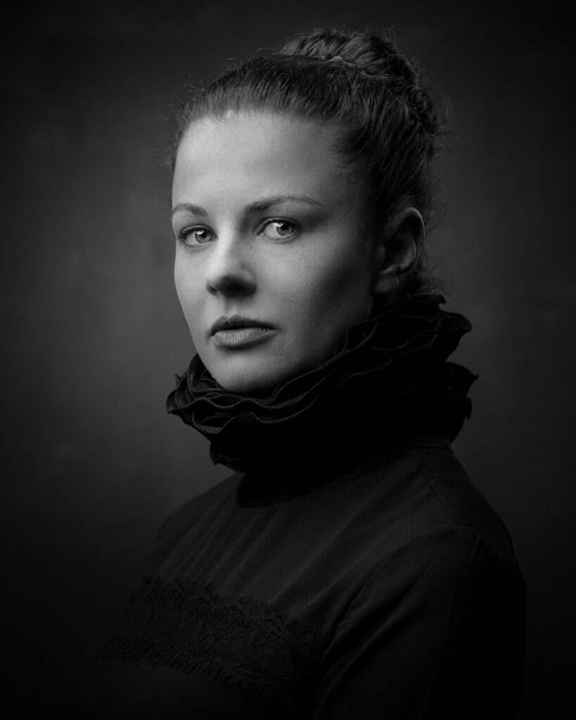 A black and white portrait of a woman with hair in a bun, wearing dark clothing with a textured collar, gazing downward against a dark background.