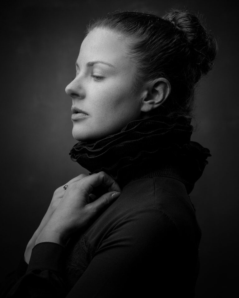 A woman in profile wearing a dark turtleneck, looking downward with a contemplative expression against a dark background.
