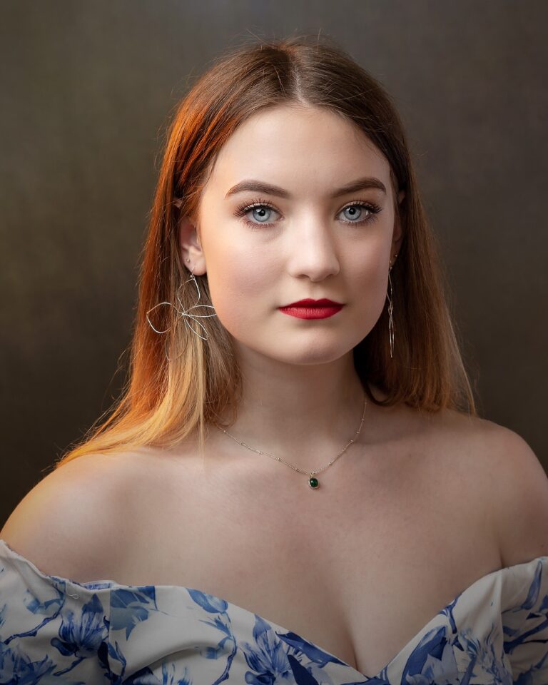 Portrait of a woman with reddish-brown hair and red lipstick, wearing hoop earrings and a blue floral off-shoulder top against a neutral background.