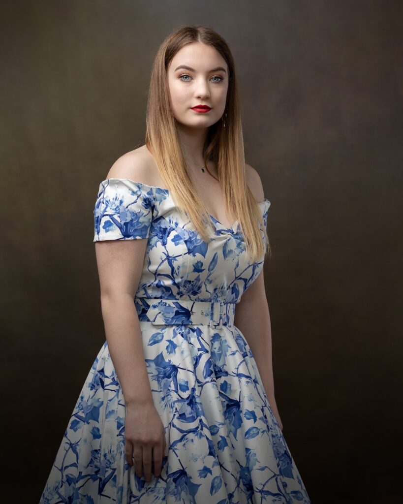 Young woman in blue and white floral off-shoulder dress with belt, standing against neutral background, looking to camera.