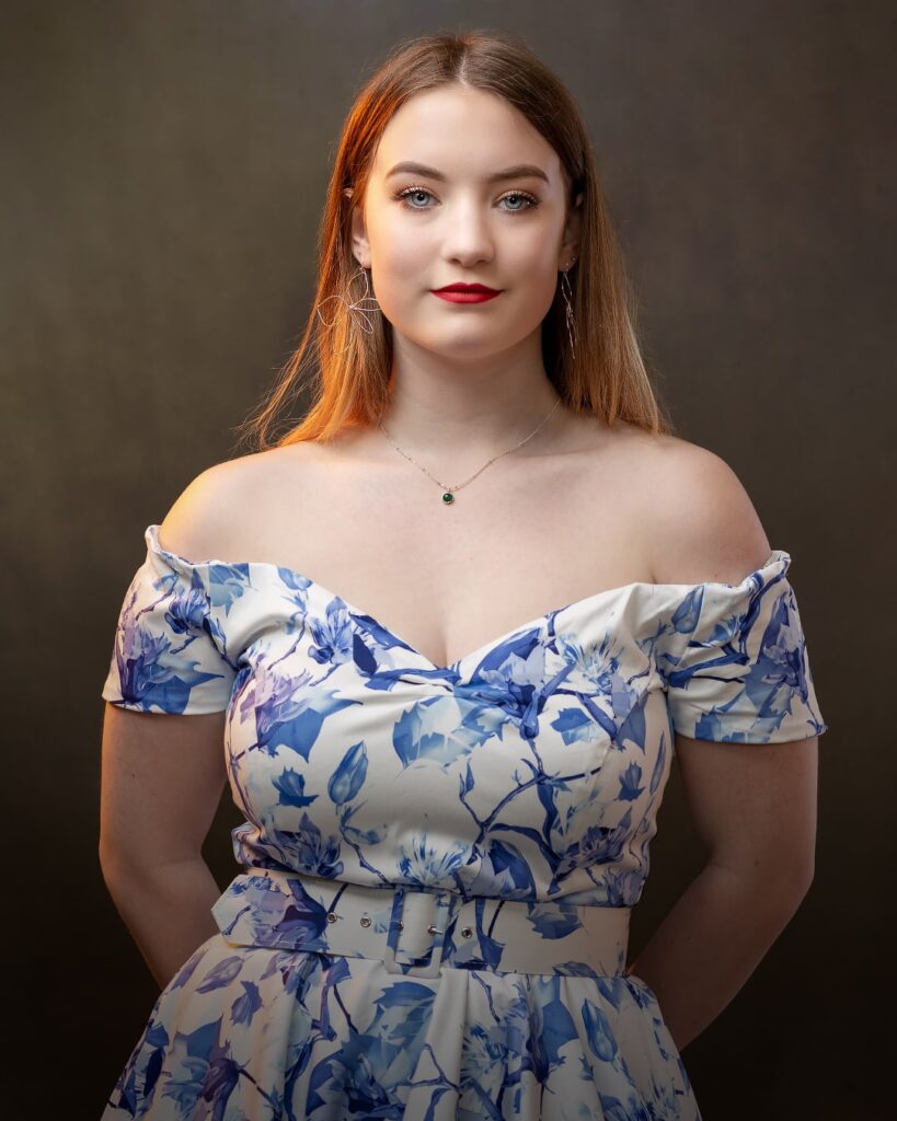 A woman with long reddish-brown hair and red lipstick wears an off-shoulder blue and white floral dress against a neutral background.