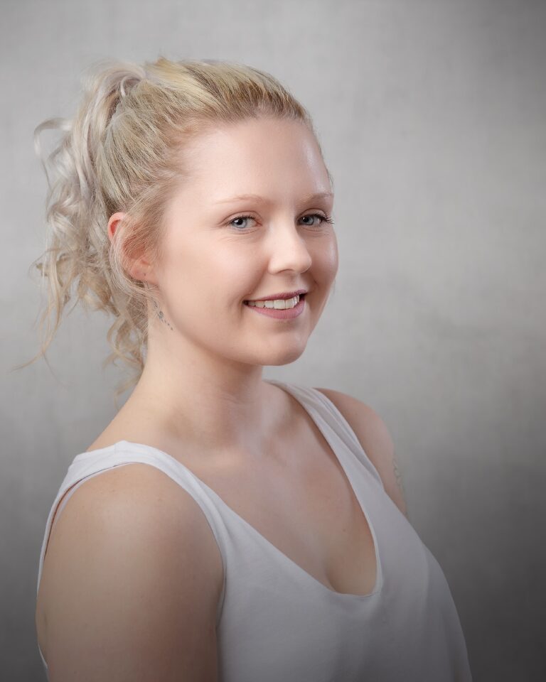 A woman with blonde hair in an updo smiles at the camera against a neutral grey background.