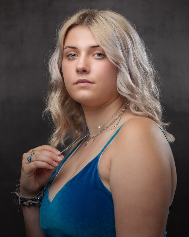 A woman with wavy blonde hair wearing a blue halter top and silver jewelry, posed against a dark grey background.