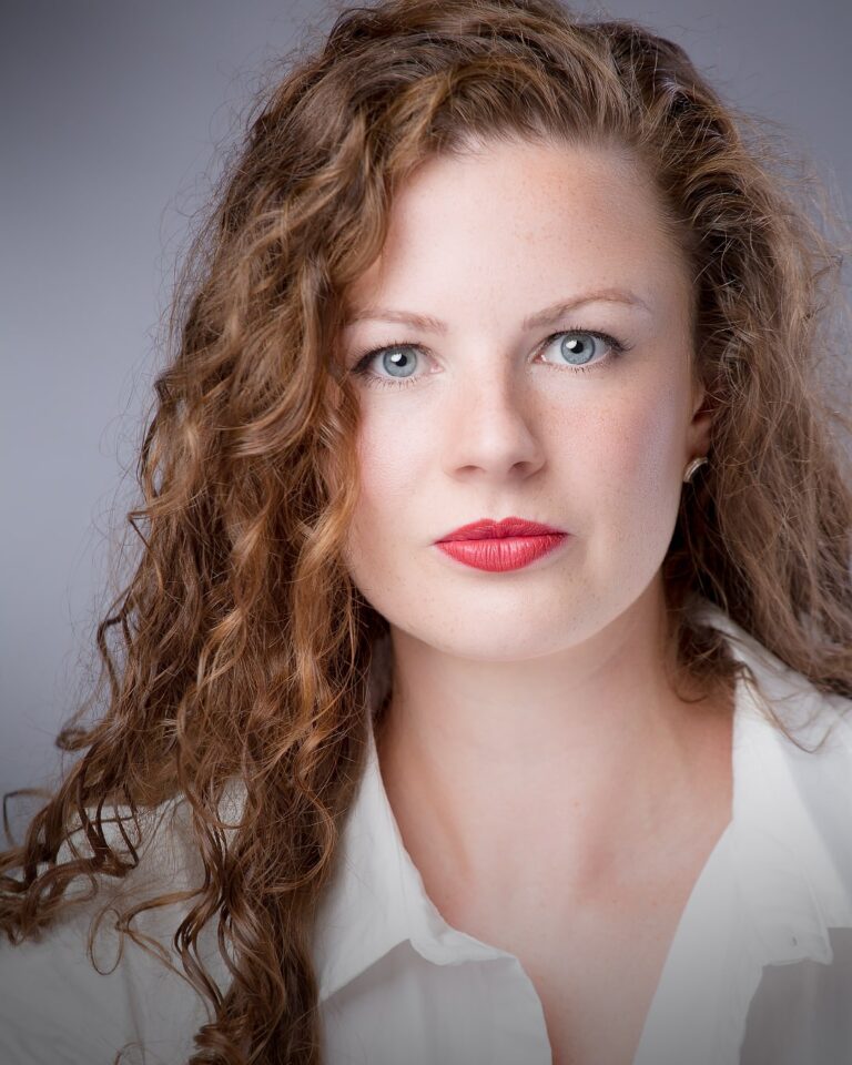 A woman with curly auburn hair and blue eyes wearing red lipstick and a white top, against a grey background.