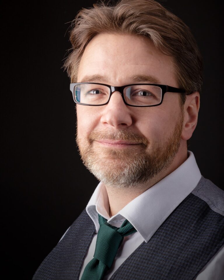 Professional headshot of a man with glasses and beard wearing a dark suit, light shirt and green tie against a black background.