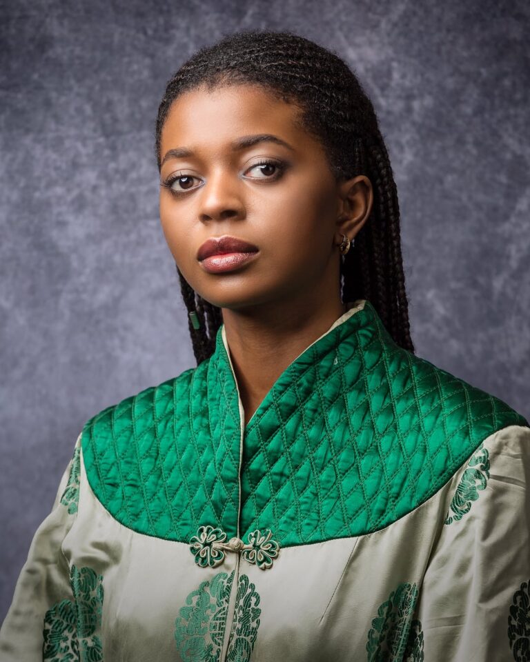 A woman in a green quilted gilet and cream tunic with green embroidery, wearing long braids and gold earrings, gazes to the side against a grey background.