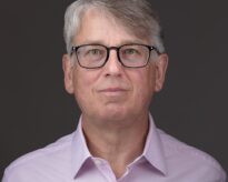 Headshot of a man with grey hair and glasses wearing a light pink shirt against a plain grey background.