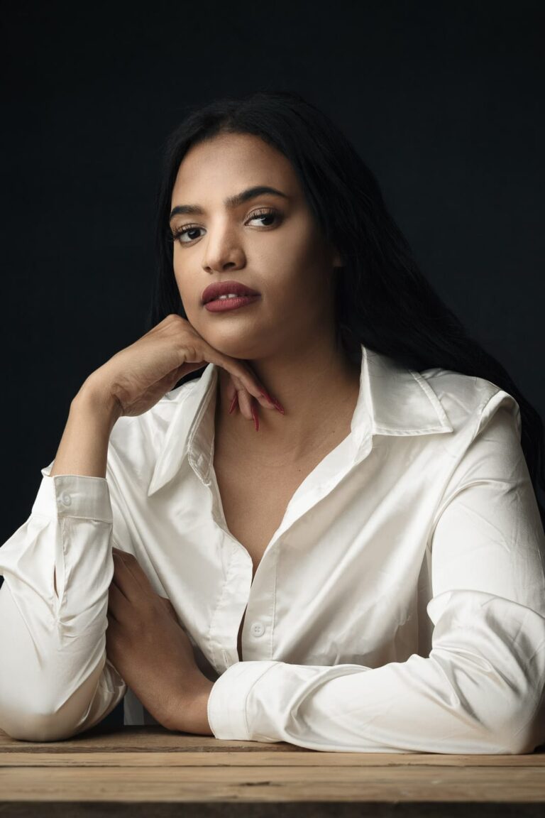 Woman with long dark hair wearing a white shirt, resting her chin on her hand, against a dark background.