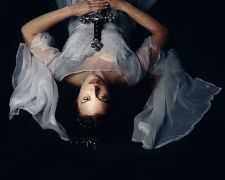 A person lies on their back against a dark background, wearing an ornate gold and studded belt, holding decorative crosses above their chest.