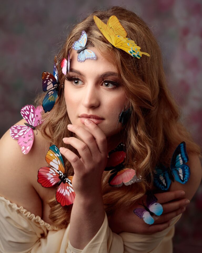 A young woman with long brown hair gazes downward, surrounded by colorful butterflies perched on her face, hair, and hand.