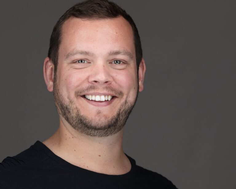 A man with dark hair and facial stubble smiles at the camera wearing a black t-shirt against a grey background.