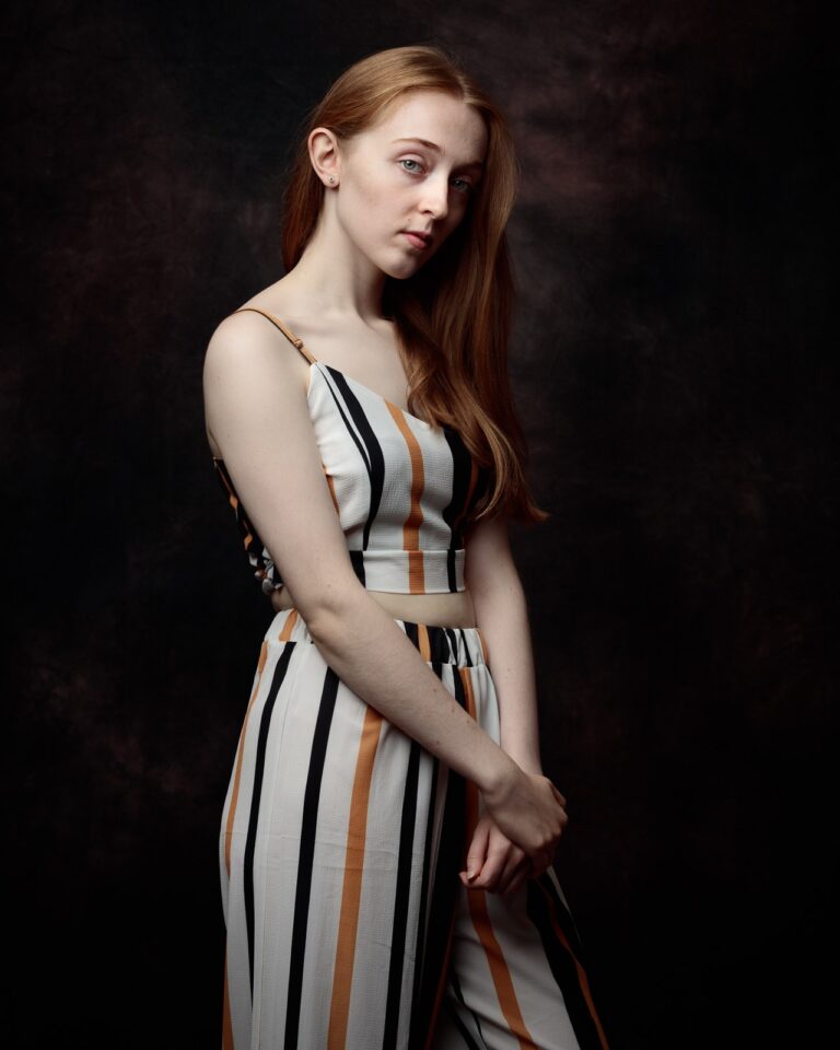 A young woman with long reddish-brown hair wearing a striped crop top and skirt stands against a dark background, looking to the side.