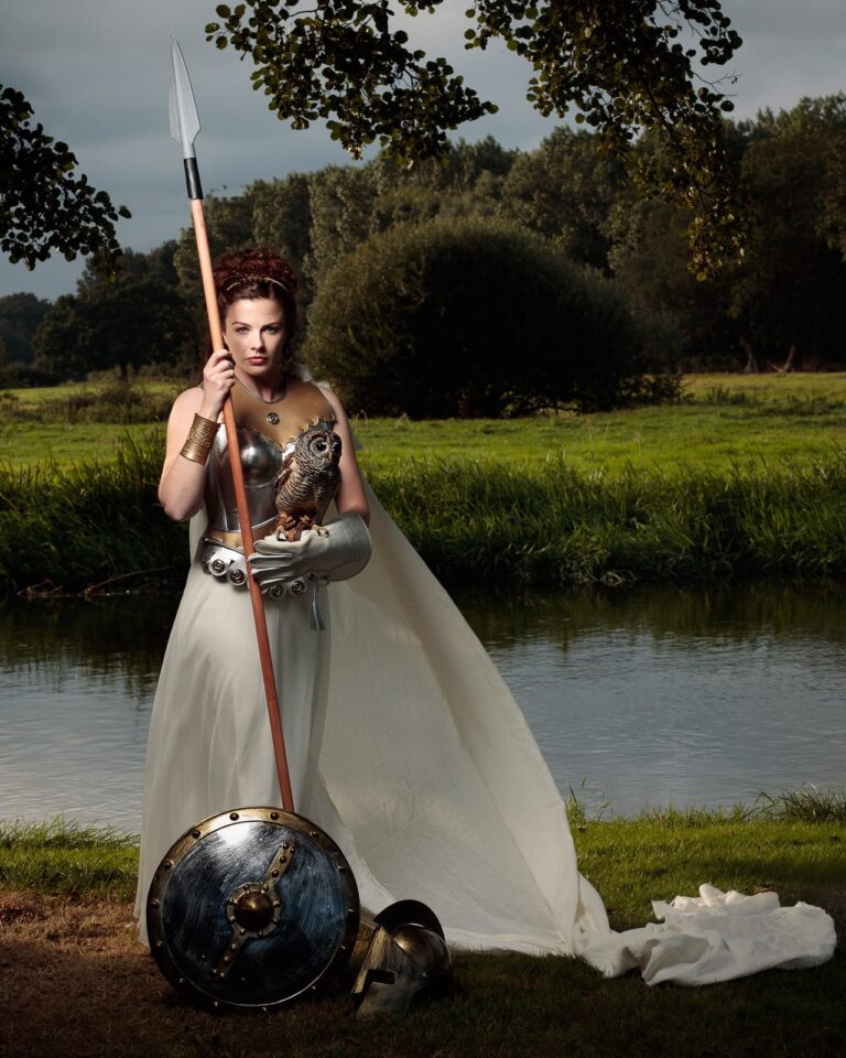 A woman in medieval warrior costume stands by a river, holding a spear and shield, wearing a bronze breastplate and white skirt.