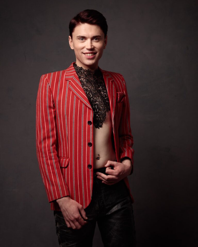 A young man smiling at the camera, wearing a red pinstriped blazer over a black lace shirt, against a dark grey background.