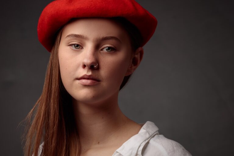 A young woman with long reddish-brown hair and a nose ring wears a bright red beret and white top against a grey background.