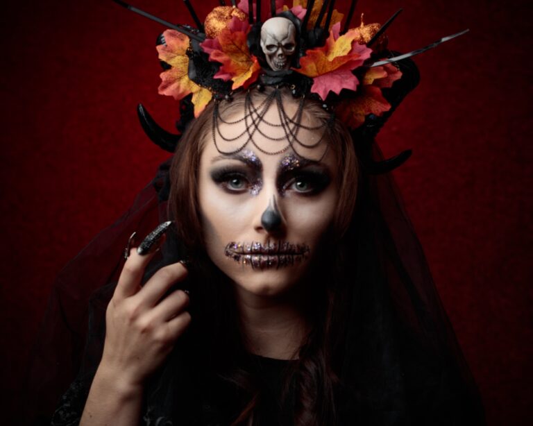 Woman with skull makeup and dark hair wearing ornate headpiece with autumn leaves and skull decoration against red background.