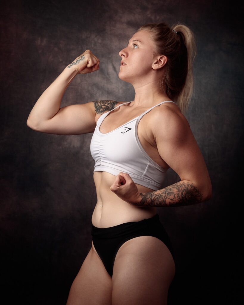 Athletic woman flexing bicep in white sports top against dark textured background, looking upward.