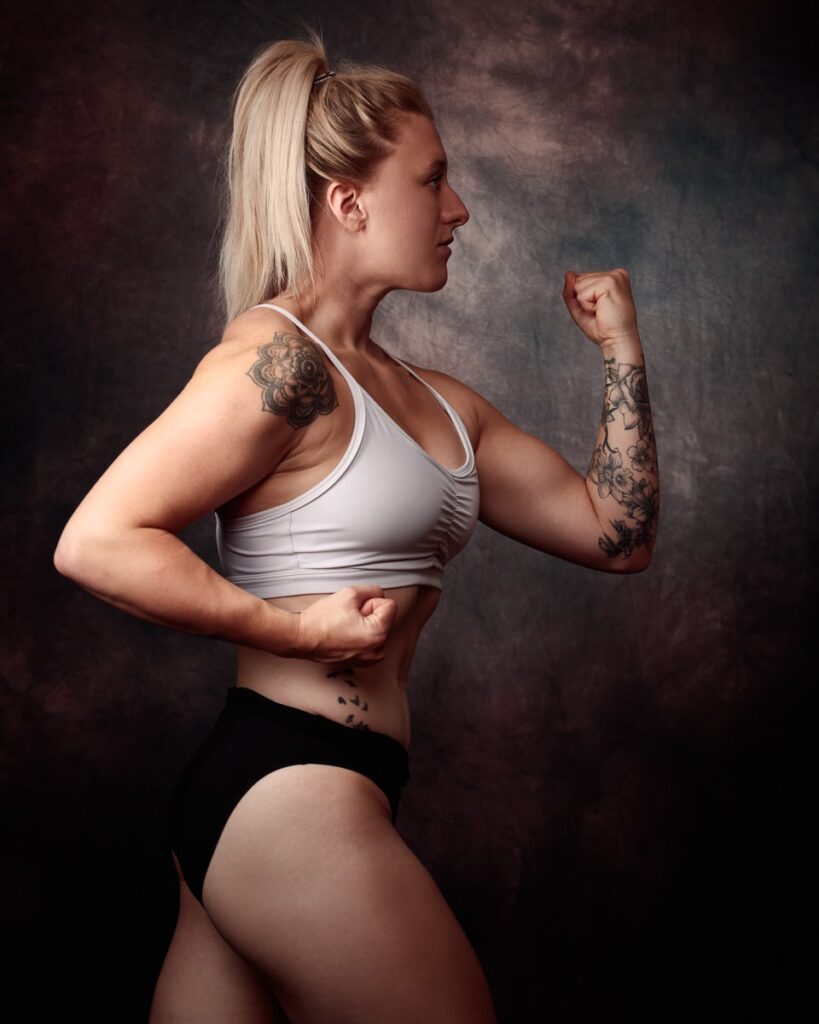 A woman in athletic wear flexes her bicep in profile against a dark textured background.