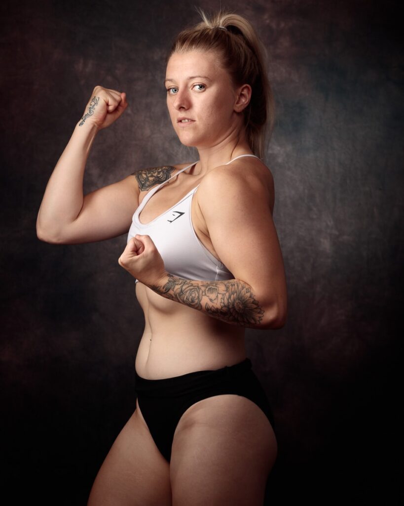 A woman in athletic wear poses against a dark background, flexing her arm in a strength pose.