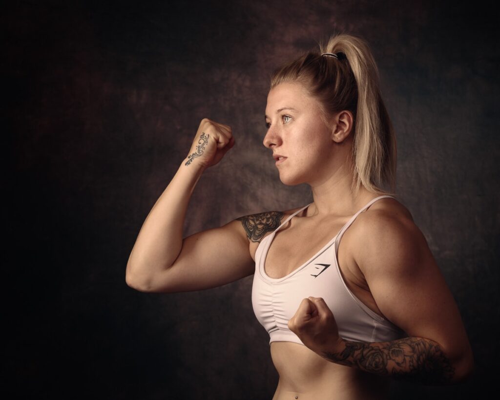 A woman in athletic wear flexes her bicep in a boxing stance against a dark background.