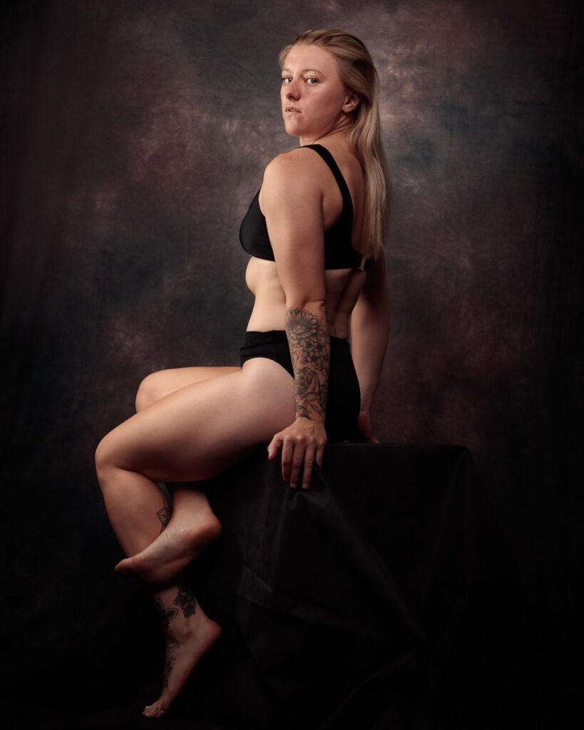 A woman with long blonde hair and visible tattoos sits on a dark surface against a textured grey-brown background, wearing black sports attire.