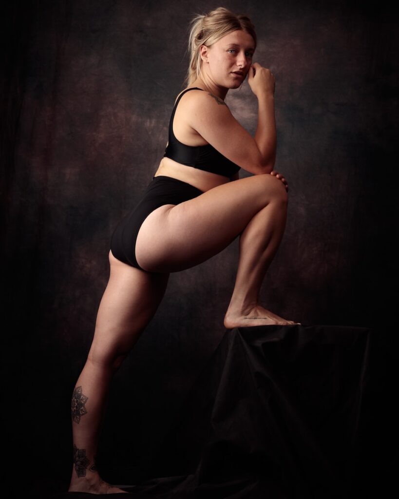 A woman in black athletic wear poses against a dark textured background, looking toward the camera with one leg raised.