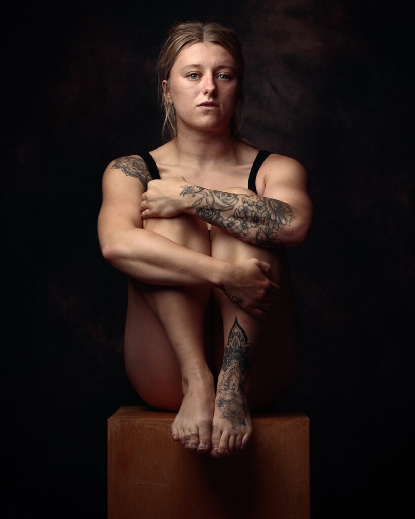 A woman with tattoos sits on a wooden cube against a dark background, arms crossed, looking directly at the camera.