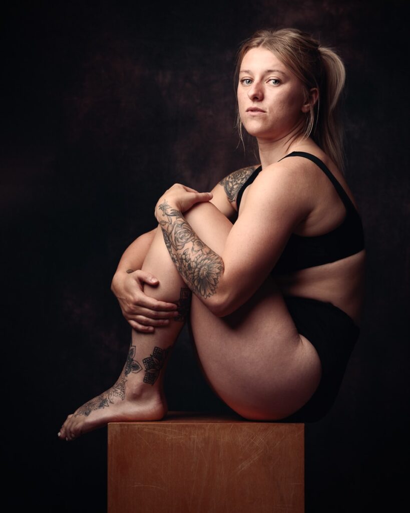 Pregnant woman with tattoos posing on wooden block against dark background in maternity portrait photograph.