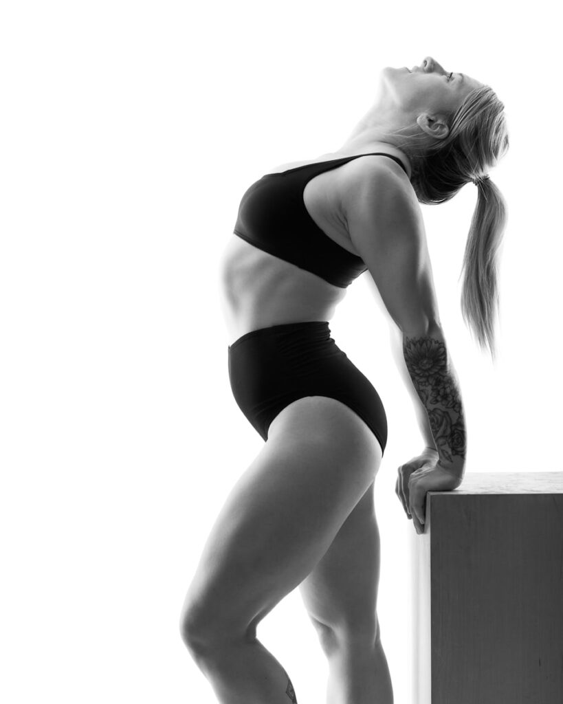 A woman in black underwear leans back on a white platform against a white background in this black and white portrait.