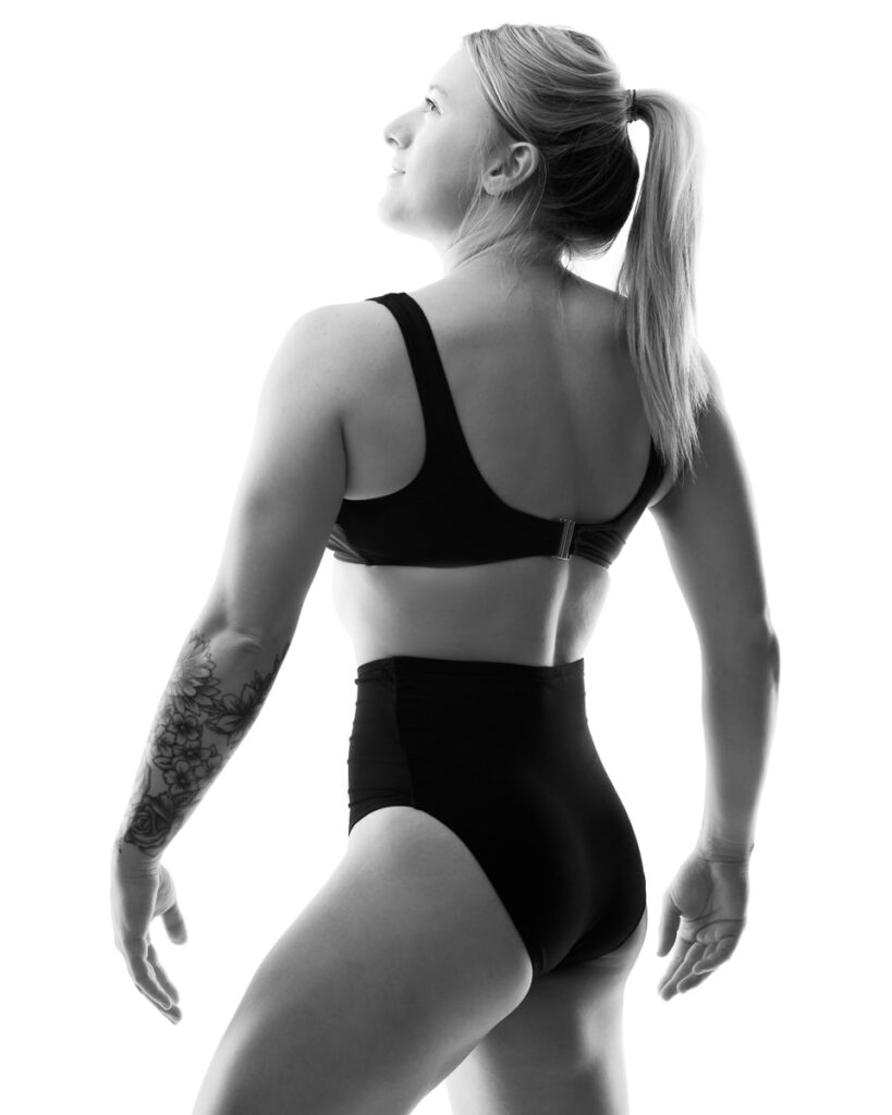 A woman in black sports bra and briefs stands in profile against a white background, looking upward.