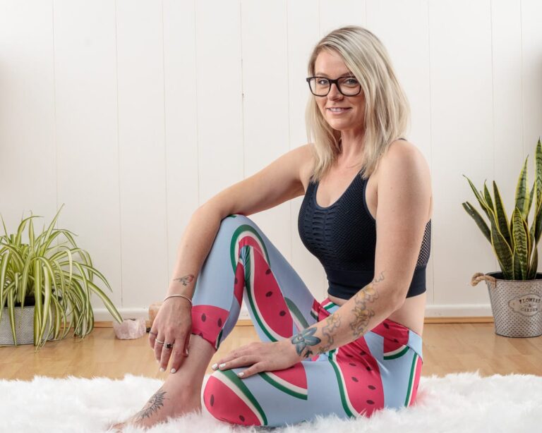 Woman sitting on white rug wearing black sports top and watermelon-print leggings, smiling at camera in bright room with plants.