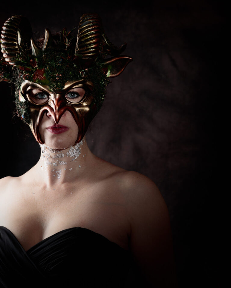 A person wearing an ornate golden mask with horns and red detailing, glasses, and glittery neck decoration against a dark background.